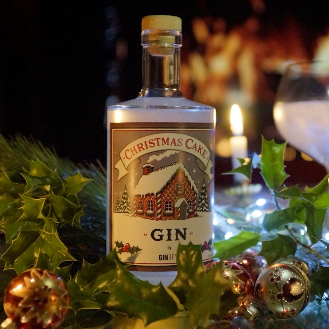 Bottle of 'Christmas Cake Gin' surrounded by holly and Christmas decorations with a blurred festive background.